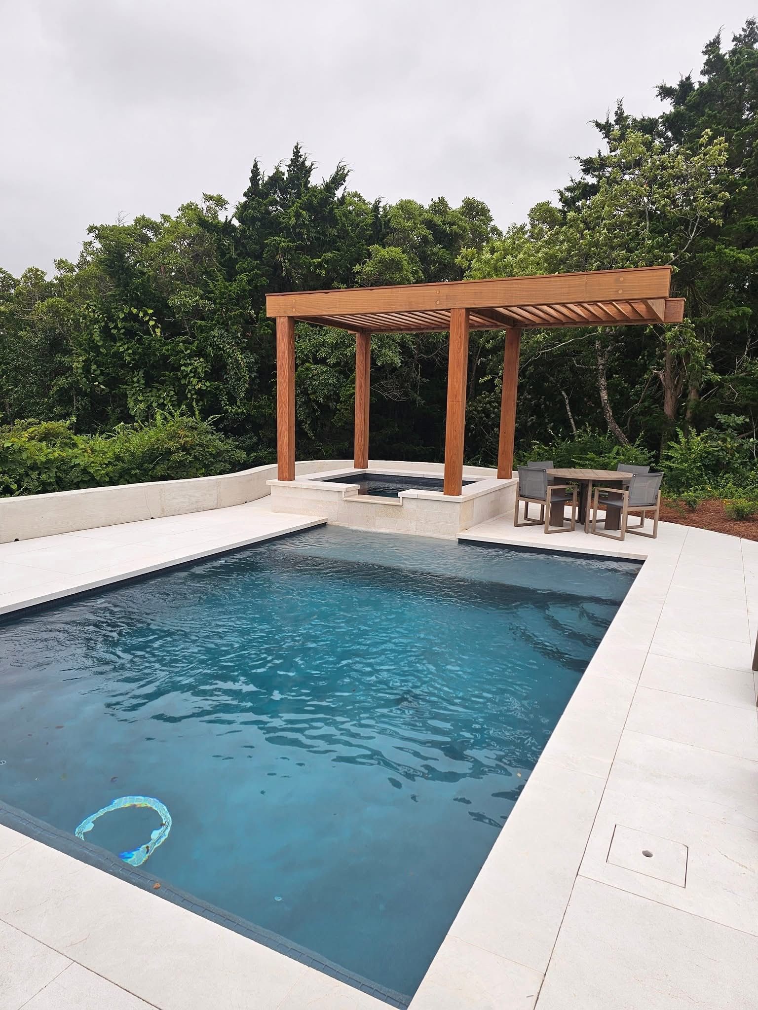 Pool with wooden pergola and surrounding trees. Turquoise water, white patio.