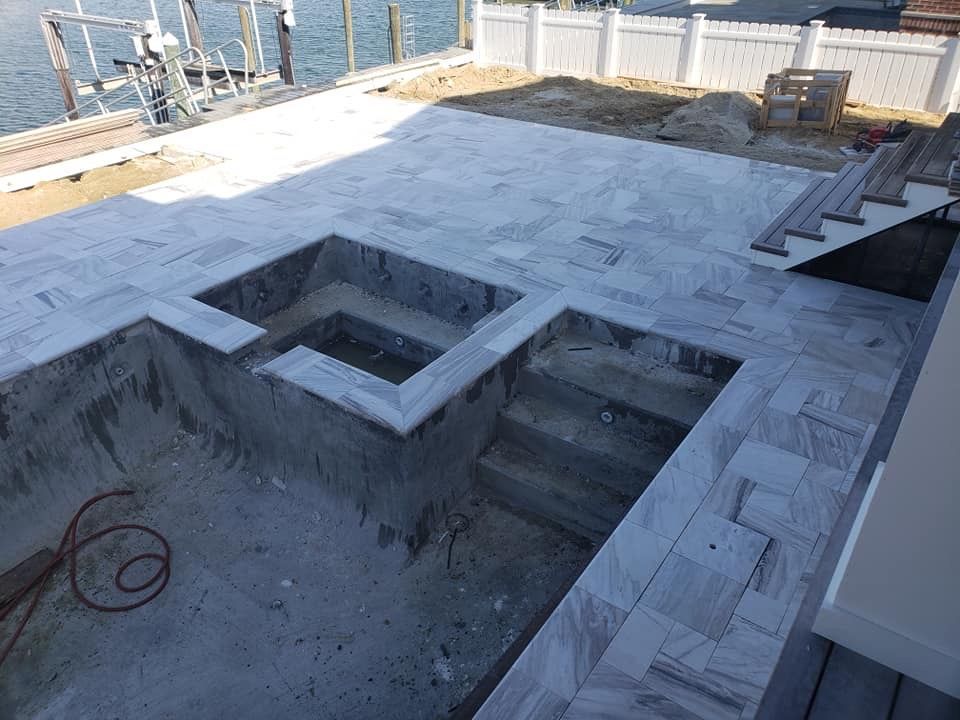 Unfinished pool with gray tile surround, steps, and a hot tub area; waterfront setting.