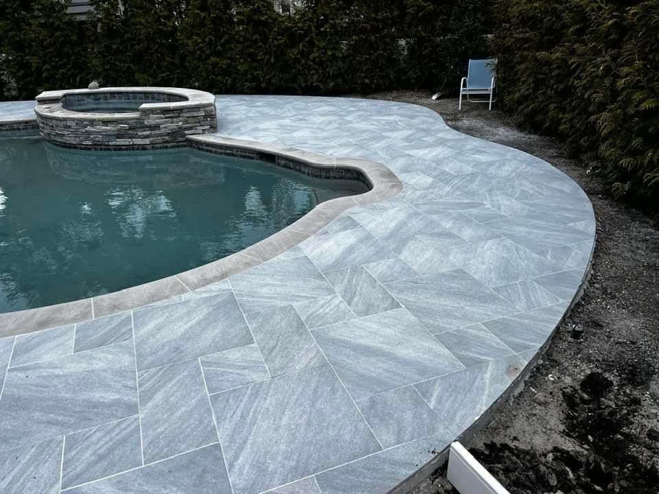 Pool with blue water and stone patio, with a hot tub and a chair.