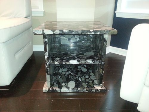 Stone side table with shelf, black and white speckled pattern, set on dark wood floor, flanked by white couches.