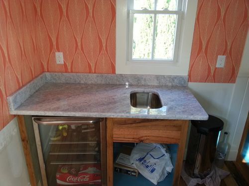 A small wet bar with a granite countertop, sink, and mini-fridge against an orange-patterned wall.