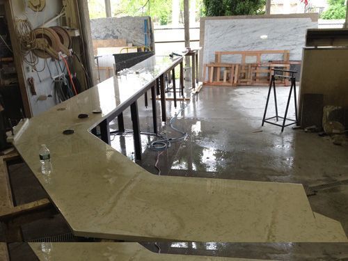 A wet, L-shaped stone countertop in a workshop; long black legs support it; granite slabs and equipment in the background.