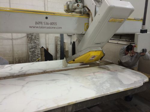 A large machine cutting a white marble slab; a worker wearing safety glasses monitors the process.