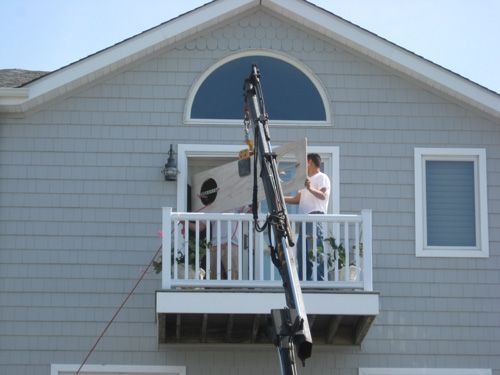 Crane lifting a door onto a balcony of a gray house. Man in white shirt directs the process.