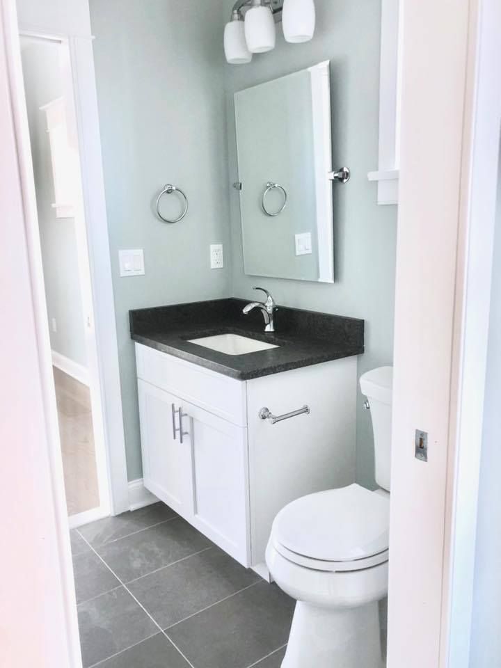 Small bathroom with white vanity, toilet, and gray tiled floor. Light blue walls and a square mirror.