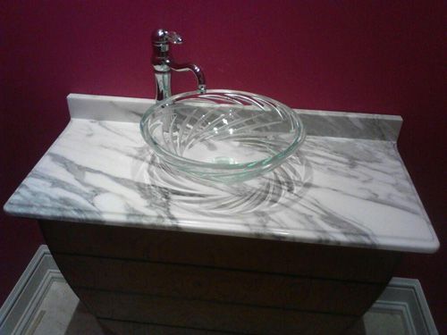 Glass bowl sink on marble counter with chrome faucet, set against a dark pink wall.
