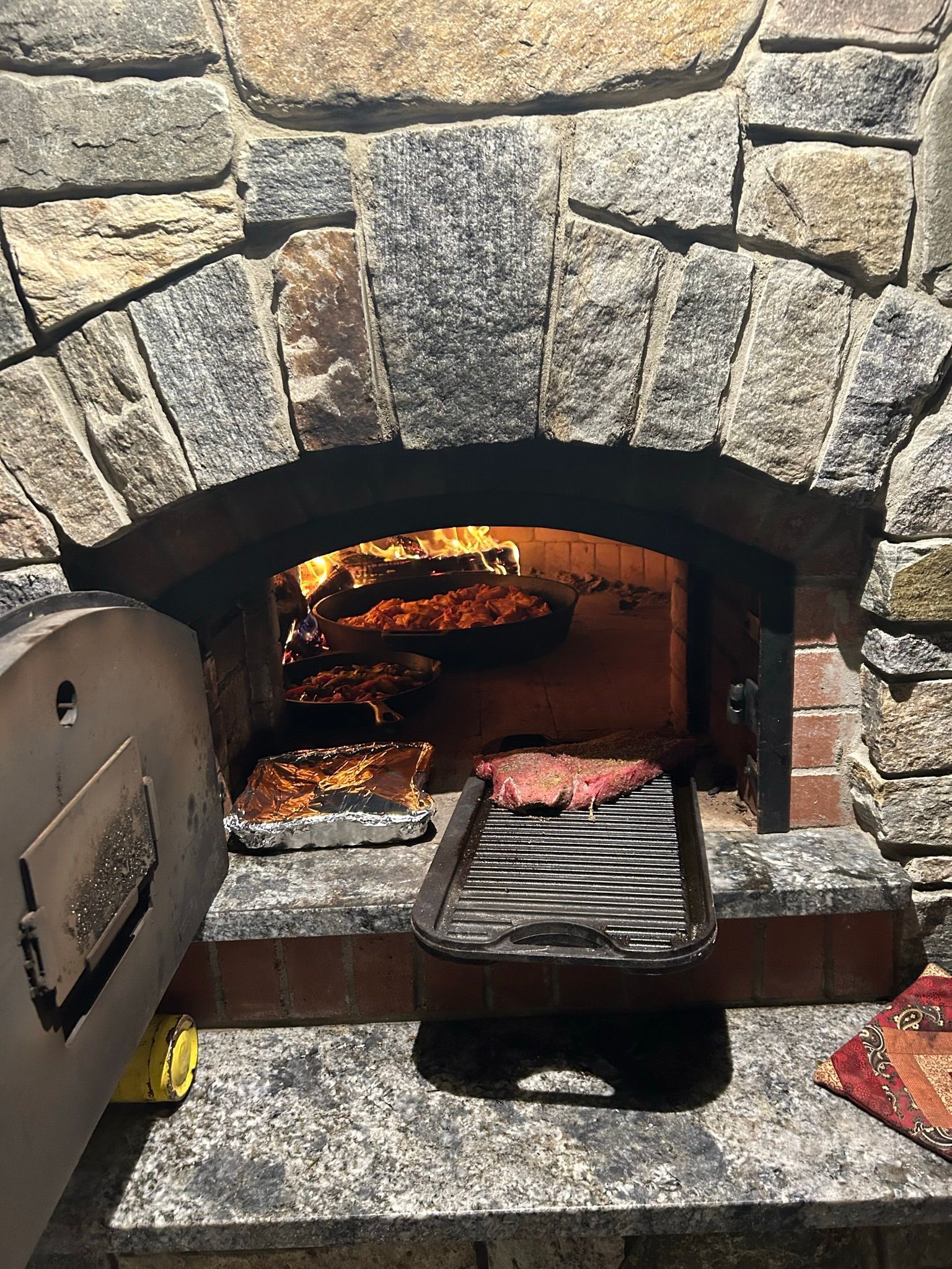 Steak grilling in a stone oven with flames visible. Oven has an arched opening and open door.