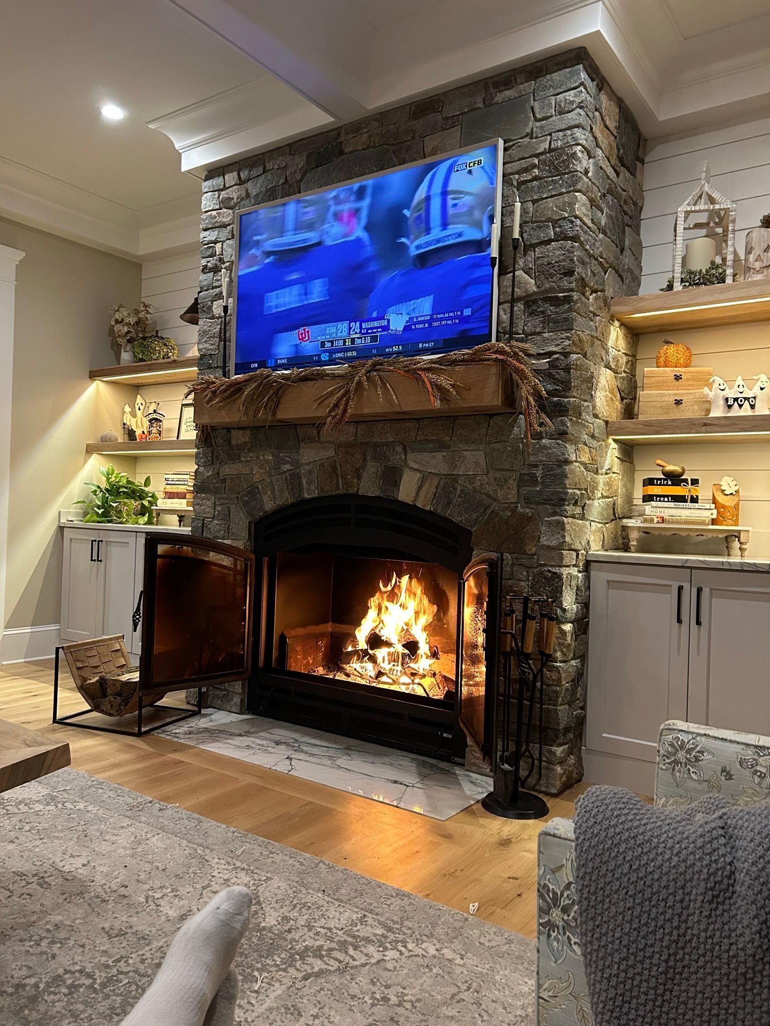 Fireplace with lit fire and mounted TV on a stone wall, shelves on either side, hardwood floor, and a rug.