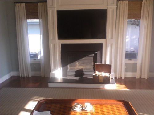 Living room with fireplace, TV, and windows, featuring white curtains, light wood floors, and a brown coffee table.