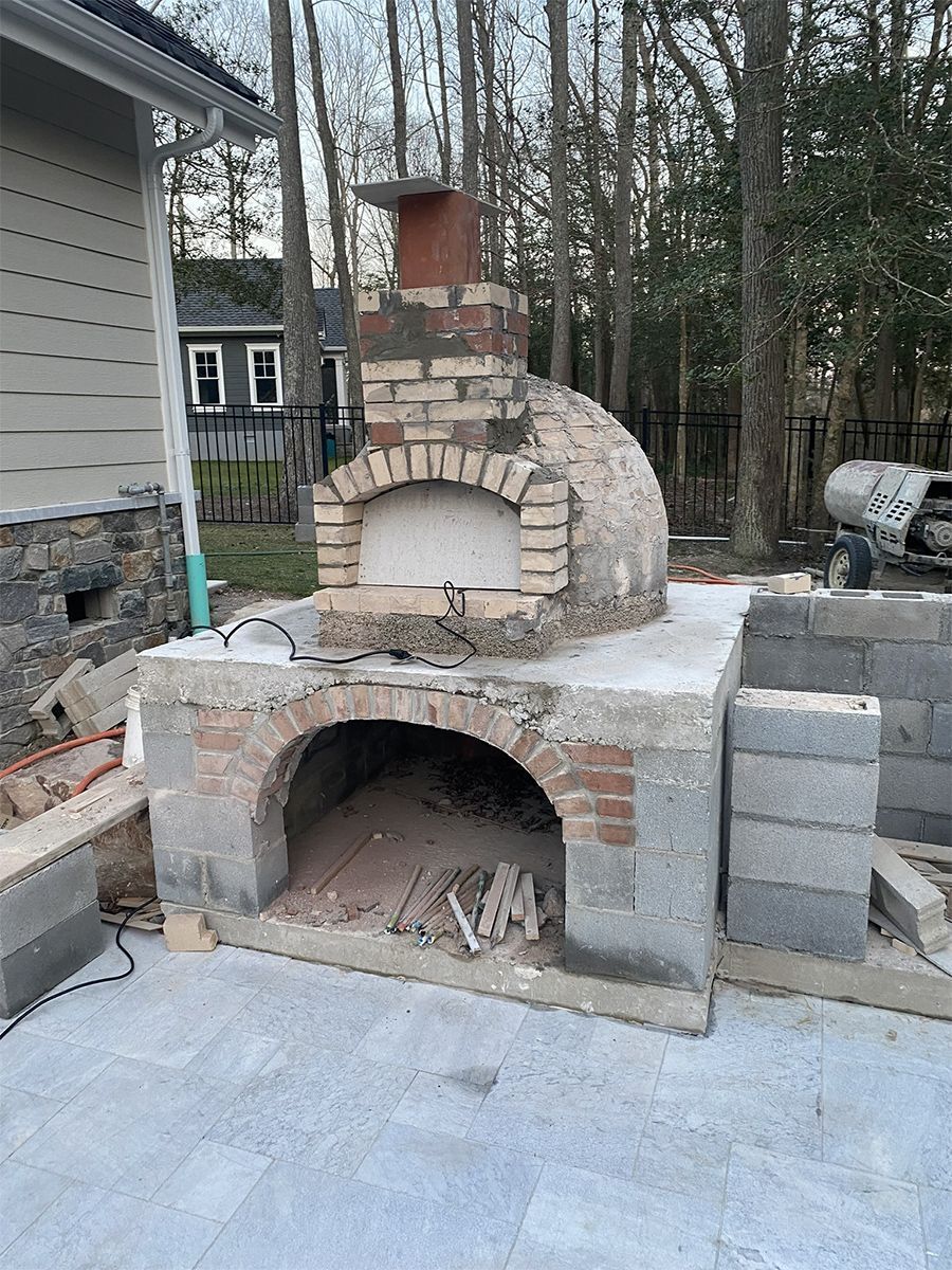 Brick pizza oven under construction outdoors, with chimney, arched opening, and concrete base.