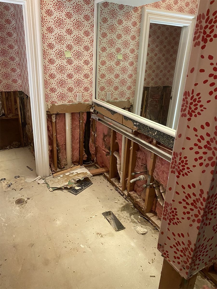 Bathroom wall under renovation, exposing studs, pink insulation, and plumbing. Red floral wallpaper. Concrete floor.