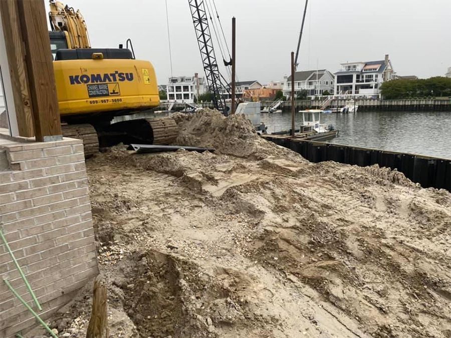 Yellow Komatsu excavator on a sandy bank, near water, with buildings in the background. Construction site.