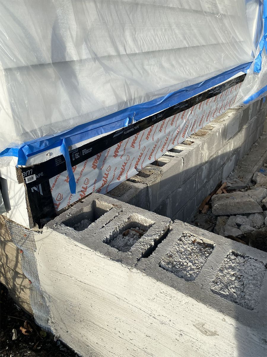 Concrete blocks partially built around a window frame, covered with protective plastic sheeting and blue tape.