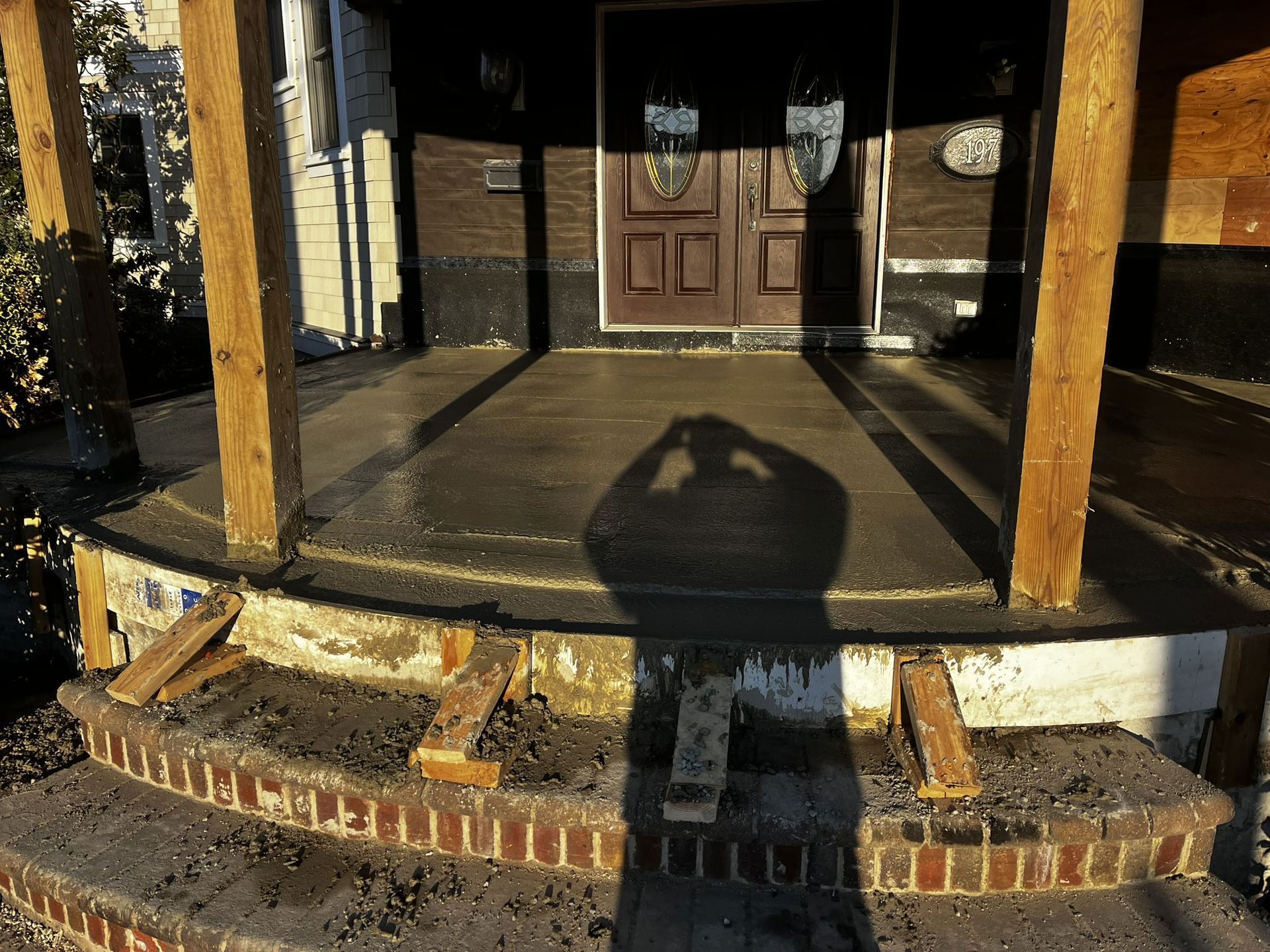 Newly poured concrete porch with brick steps, wooden posts, and shadow of a person taking a photo.