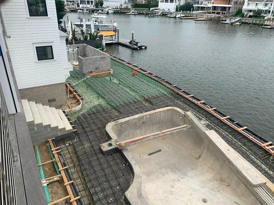 Construction site by water: Pool frame, rebar grids, green mesh, steps, boats, and houses.