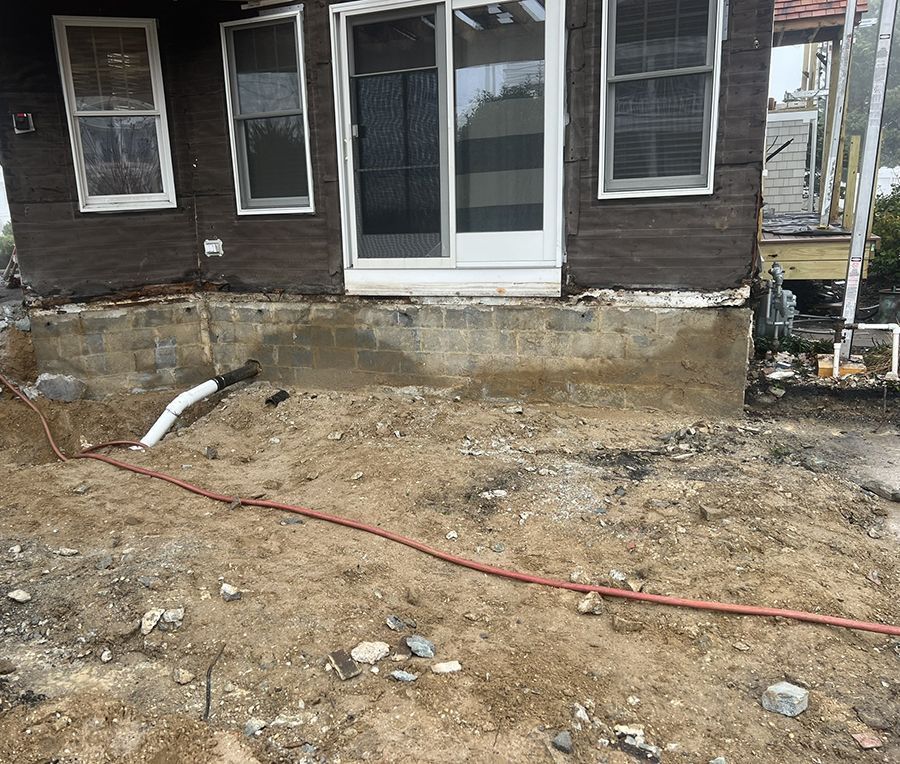 Brown house with damaged foundation; a red hose and white pipe are on the dirt ground.