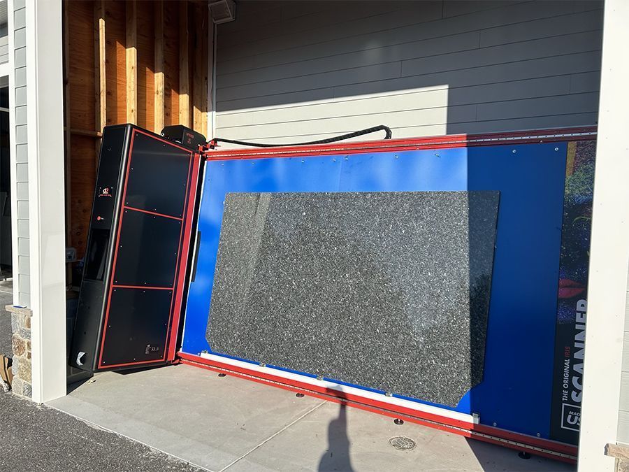 Large scanning device with a dark granite slab inside, next to a black machine, leaning against a wooden wall outdoors.