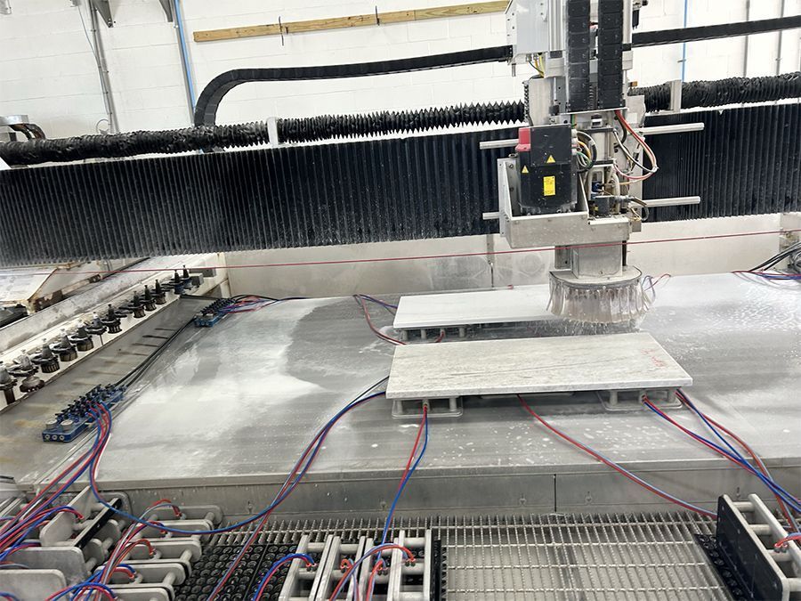CNC machine cutting a gray stone slab, with water spray and electrical wiring visible.