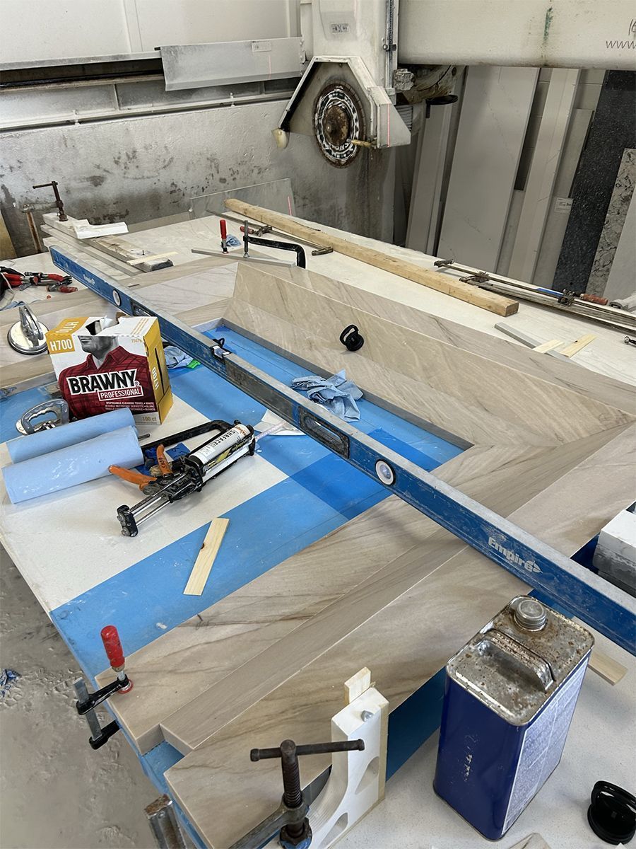 Herringbone patterned stone being worked on a table, with level, tools, and a large saw in background.