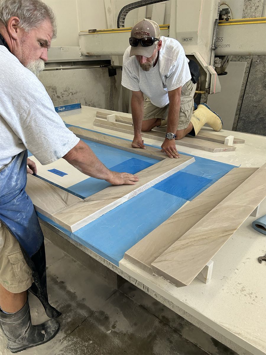 Two men working on a countertop. One uses a level, other aligns wood pieces on blue material. Workshop setting.