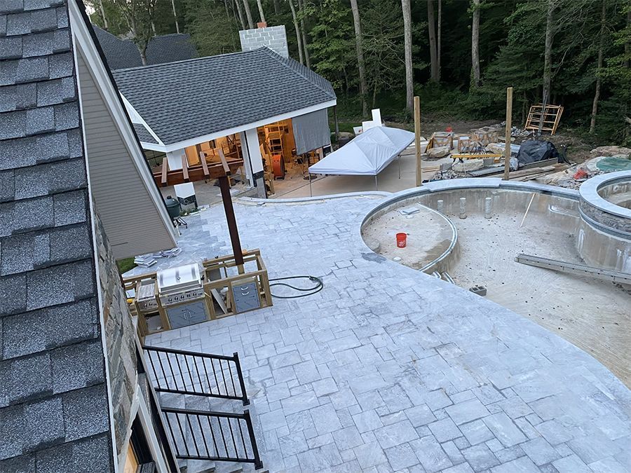Patio with gray stamped concrete, unfinished pool, building with gray roof, forest backdrop.