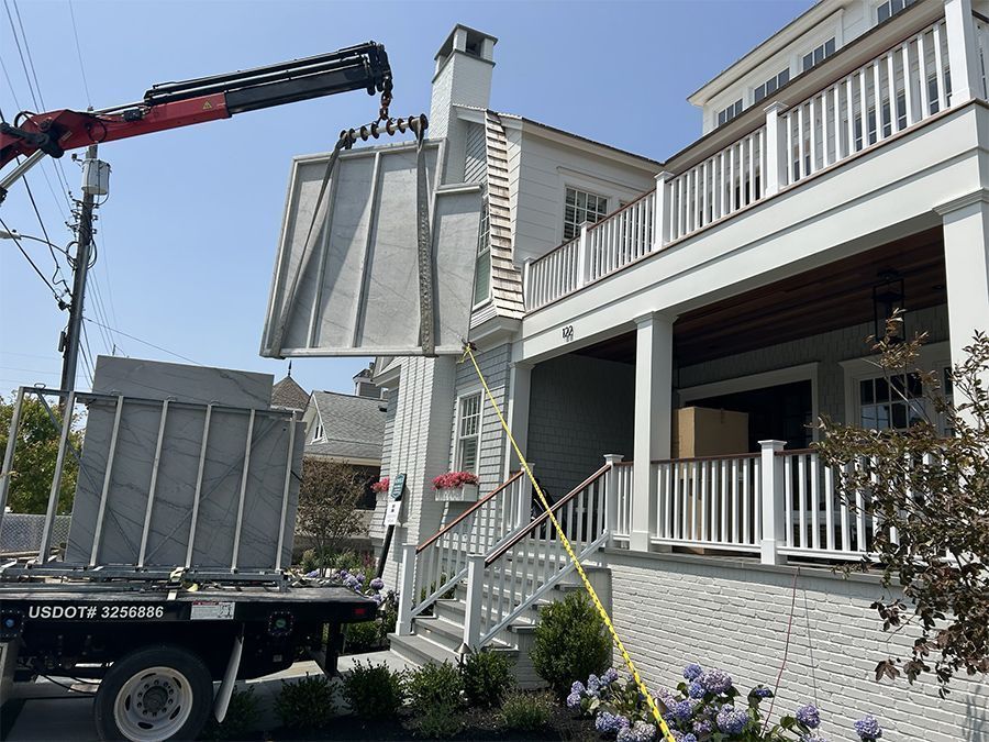 A crane lifts a large concrete panel near a white house with a porch and steps. Blue sky.
