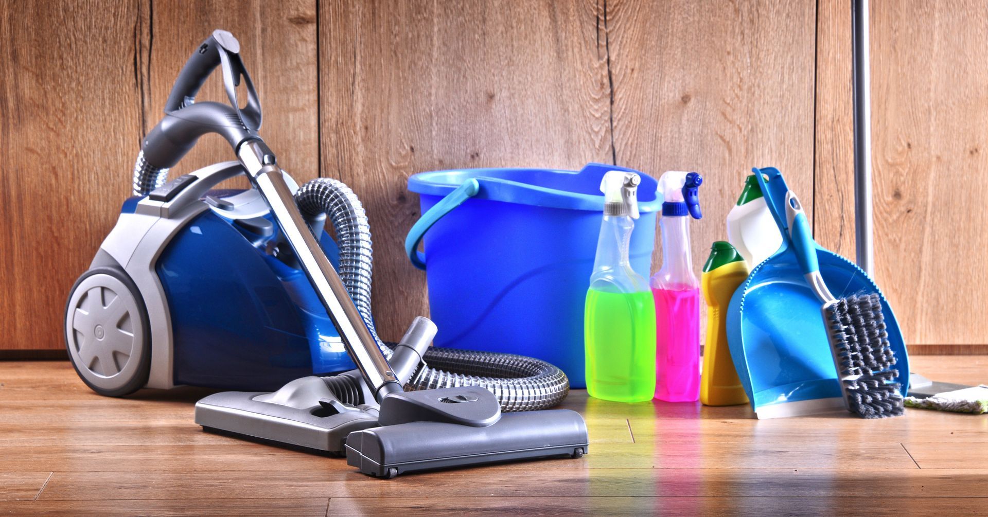 Cleaning supplies on wood floor: vacuum, bucket, spray bottles, and brush.