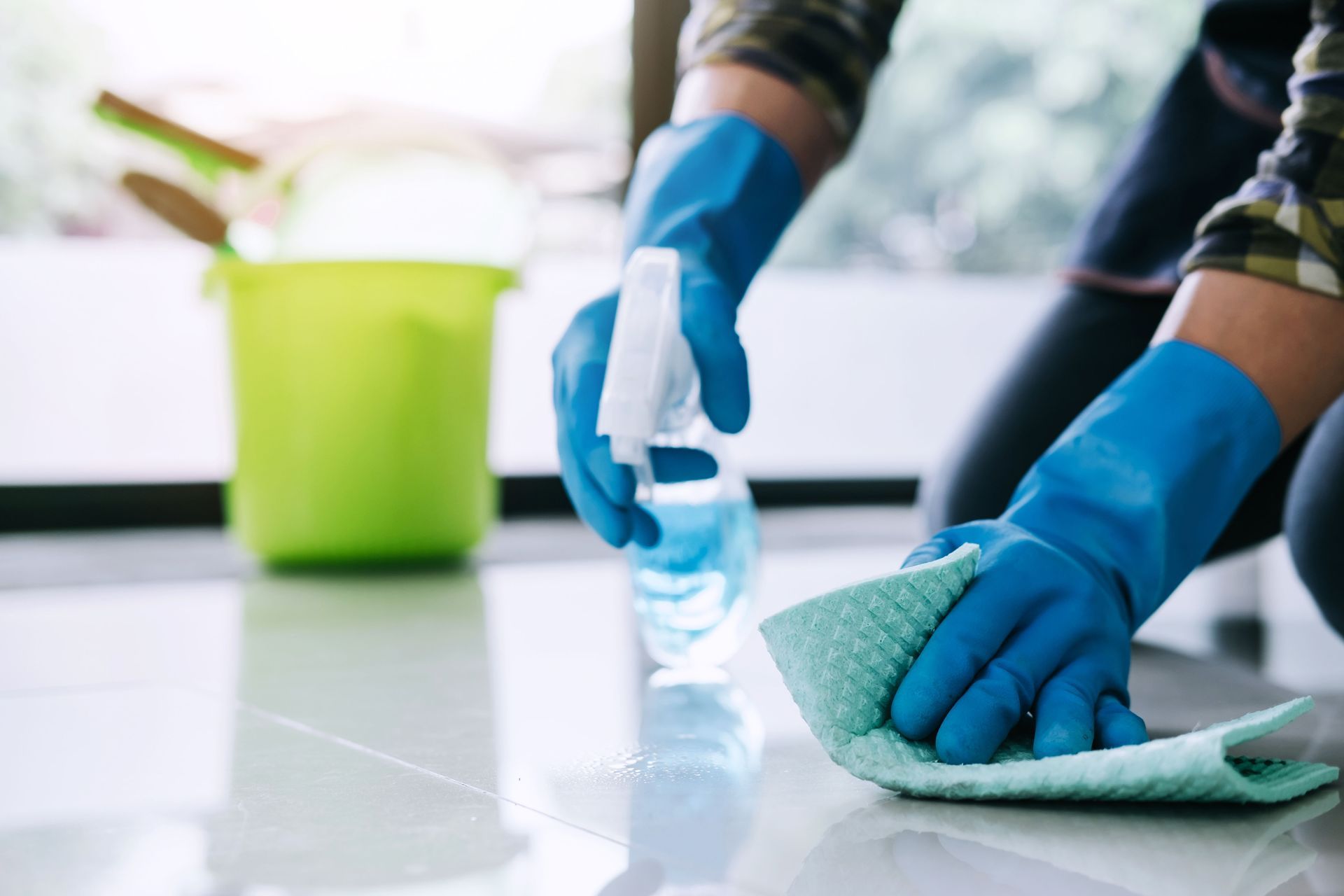 Person wearing blue gloves cleans a white tiled floor with a spray bottle and cloth; green bucket in the background.