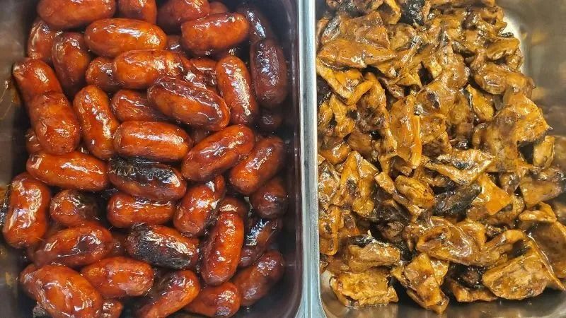 Two metal trays holding cooked food: glazed sausages and sauteed mushrooms.