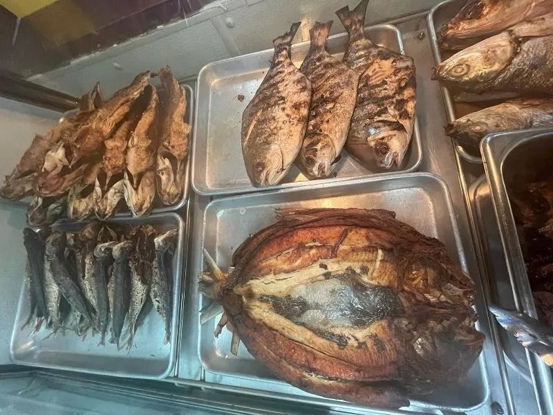 Display of cooked fish in a restaurant setting. Varieties include whole grilled and smoked fish in metal trays.