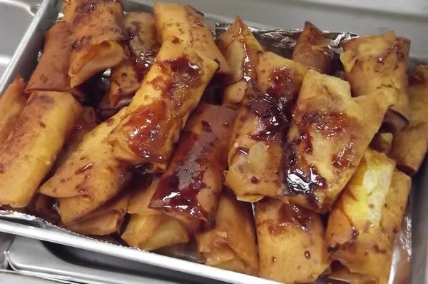 Fried lumpia, drizzled with sauce, piled in a metal tray.