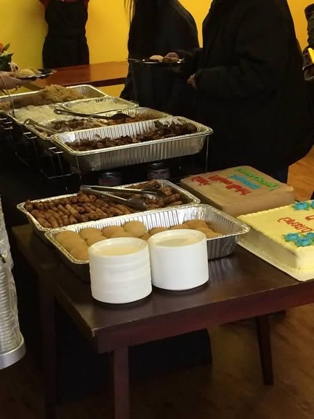 Food buffet with various dishes in aluminum trays, two cakes, and serving utensils.