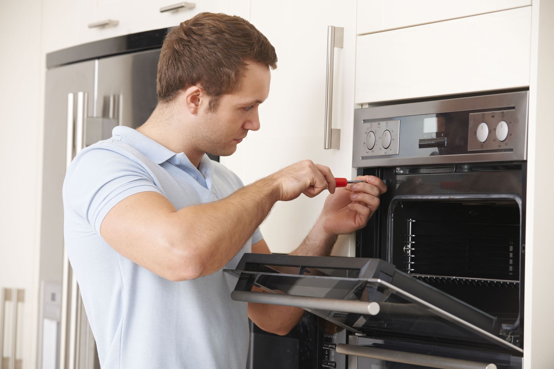 A person wearing a light blue polo shirt uses a screwdriver to repair a stainless steel oven in a kitchen.