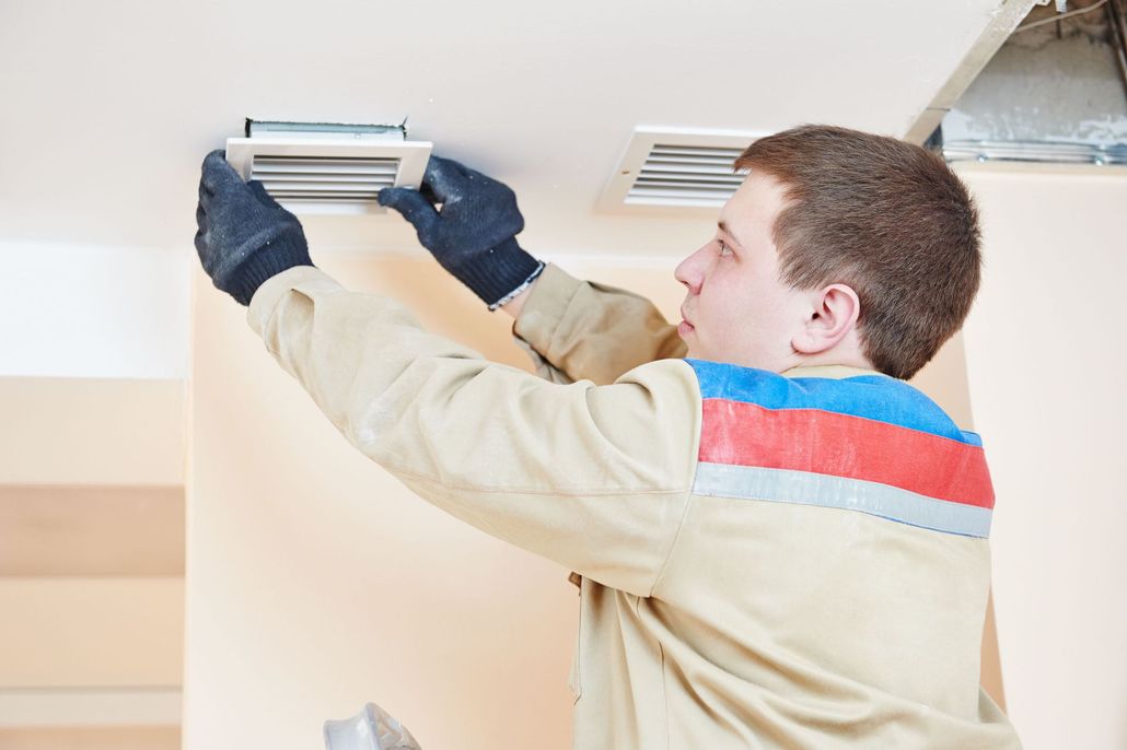 A worker in a tan uniform and black gloves installs a white ventilation grill into a ceiling.