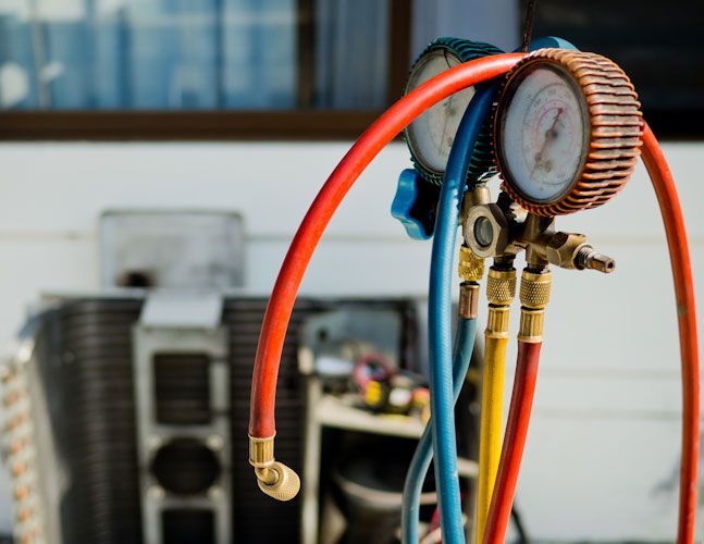 An HVAC manifold gauge set with red, blue, and yellow hoses positioned in front of an outdoor air conditioning unit.