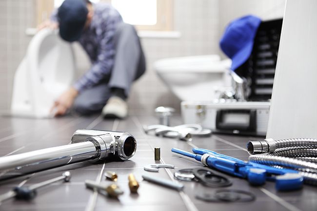 A plumber kneels on a tiled floor working on bathroom fixtures, with various tools scattered in the foreground.