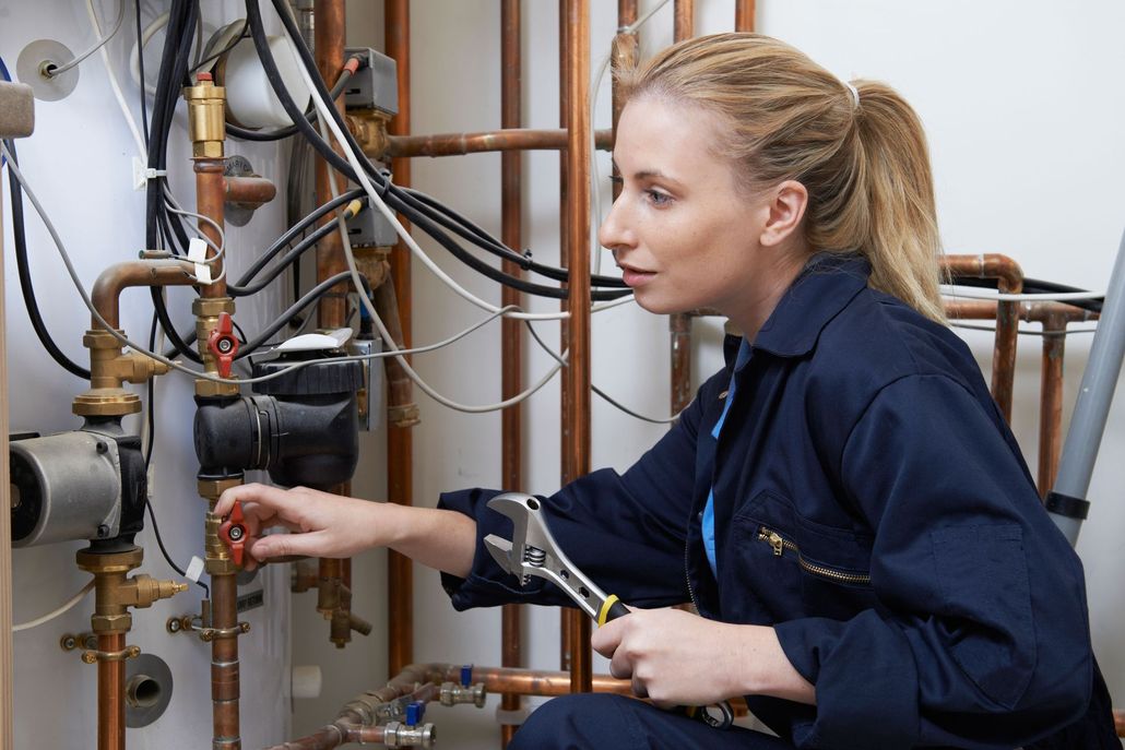 A worker in a blue uniform inspecting pipes and electrical components while holding an adjustable wrench.