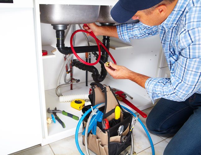 A plumber in a plaid shirt kneels under a kitchen sink, holding a pipe fitting next to a tool bag and scattered supplies.