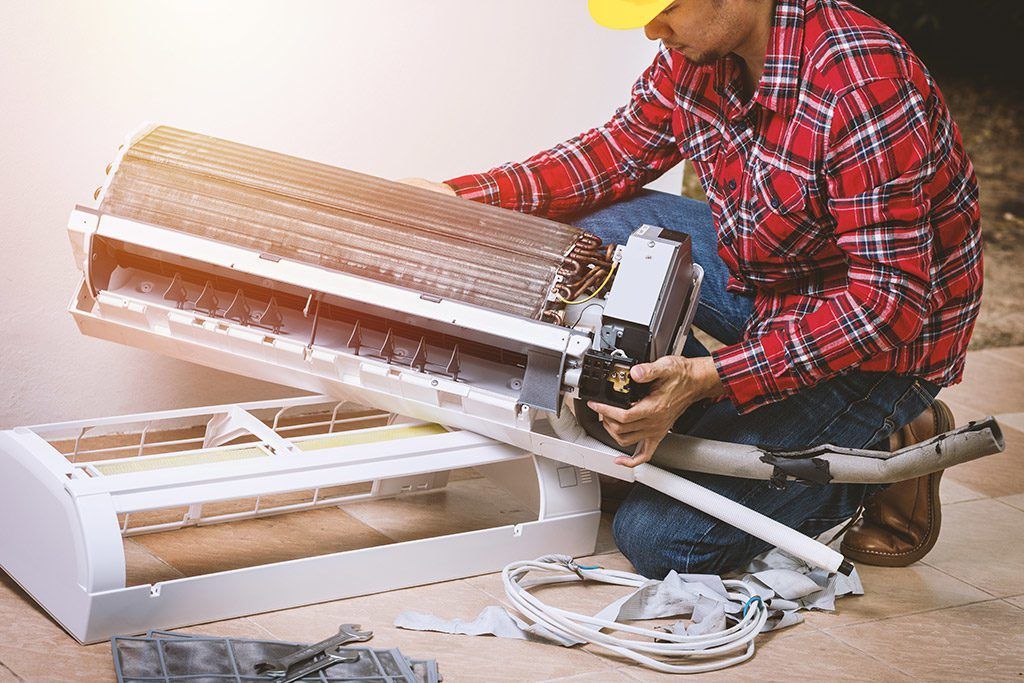 A technician in a red plaid shirt and yellow hard hat repairs a wall-mounted air conditioning unit.