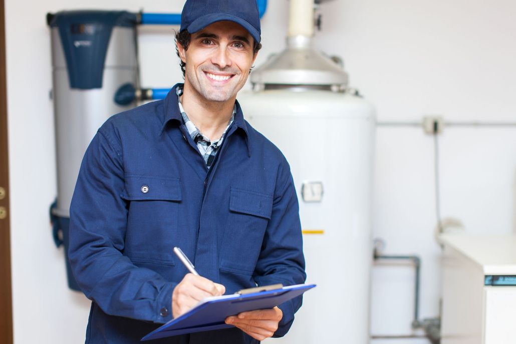 A worker in a blue uniform and cap smiles while writing on a clipboard in front of a water heater in a utility room.