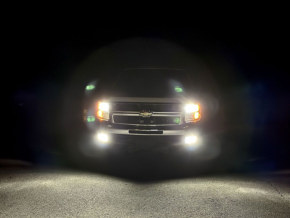 Black truck with headlights and fog lights on at night, illuminating a dark road.