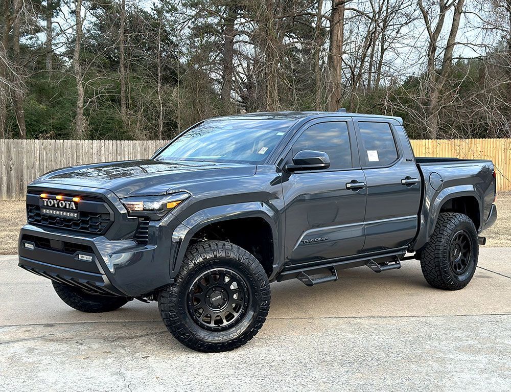 Dark gray Toyota Tacoma truck with black wheels and off-road tires parked outside.