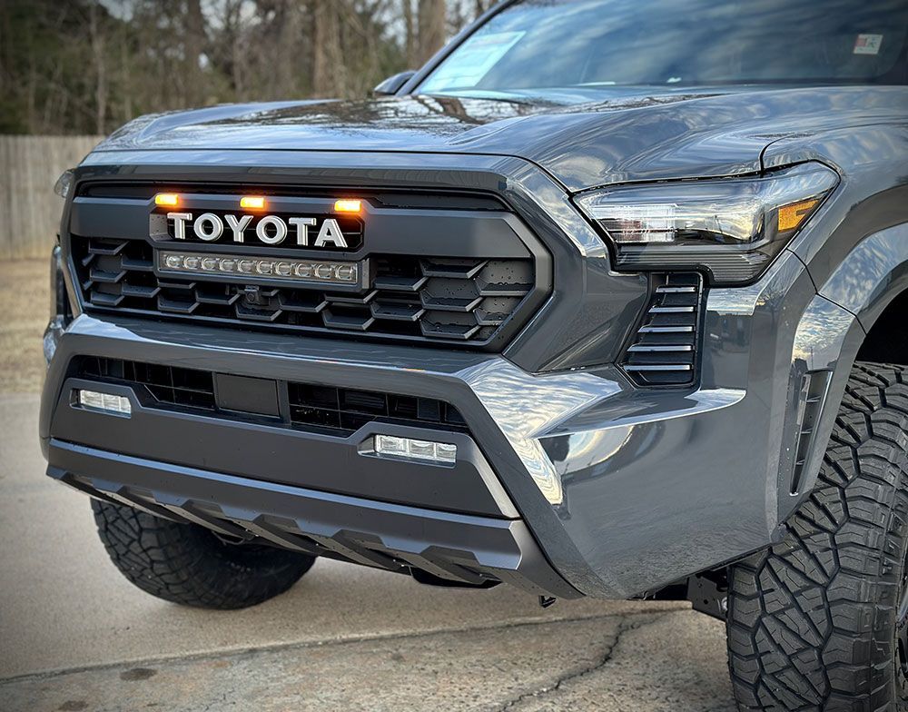Gray Toyota Tacoma truck with modified black grill, LED lights, and off-road tires.