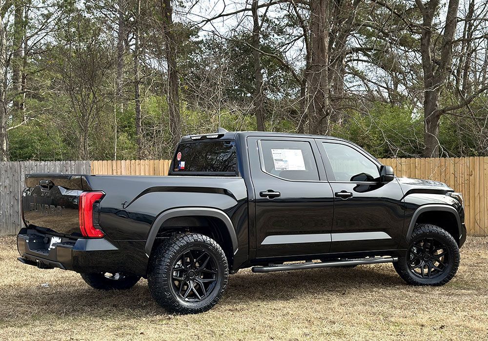 Black Toyota Tundra pickup truck parked on grass, with black wheels, and a wooden fence background.