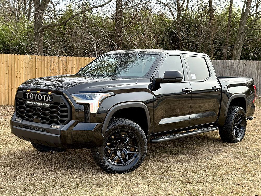 Black Toyota Tundra truck parked outside, black wheels, against a wooden fence and trees.