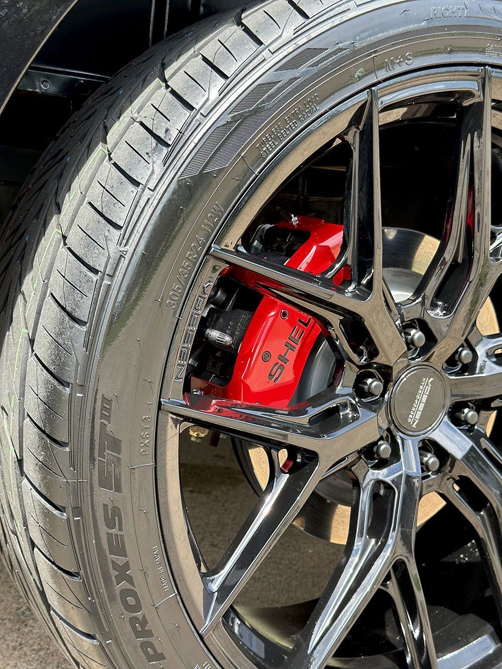 Black alloy wheel with red brake calipers and a black tire; sunlight on the tire.
