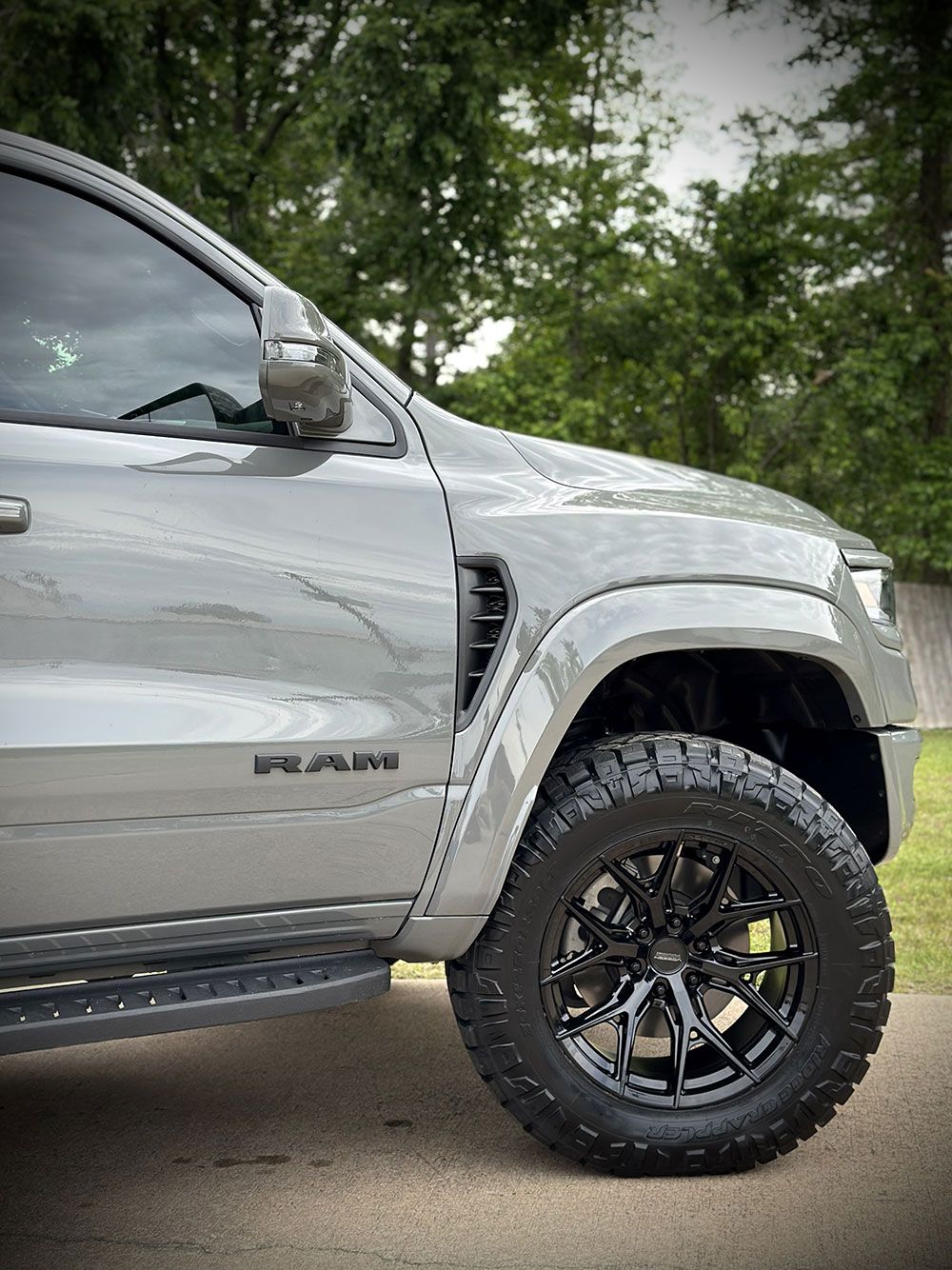 Gray Ram truck's front side, black wheel, large tire, vents, side step, outdoors.