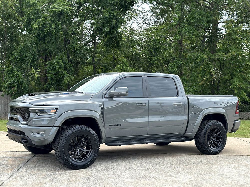 Gray Ram TRX pickup truck parked on pavement with black wheels, set against a backdrop of green trees.