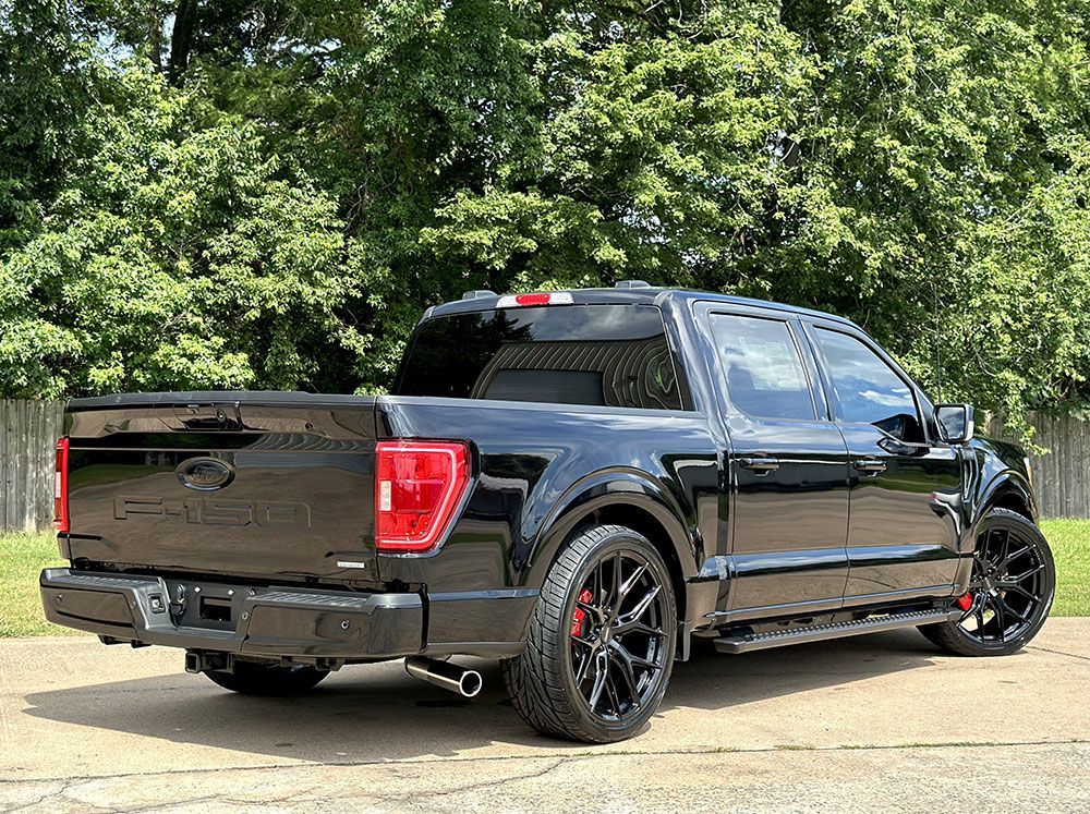 Black Ford F-150 pickup truck with custom modifications, parked on a paved surface, trees in the background.