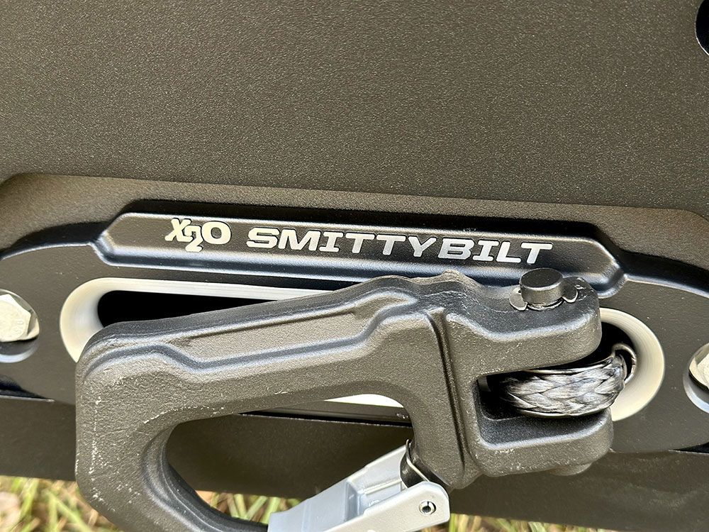 Close-up of a black Smittybilt winch with a black carabiner attached. The winch is mounted on a vehicle.